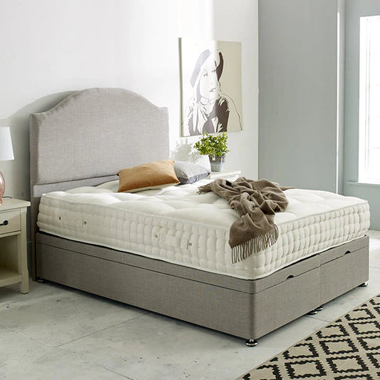 A Sencilla Divan bed base in silver plush velvet with a padded headboard, displayed in a bedroom setting with ottoman storage visible